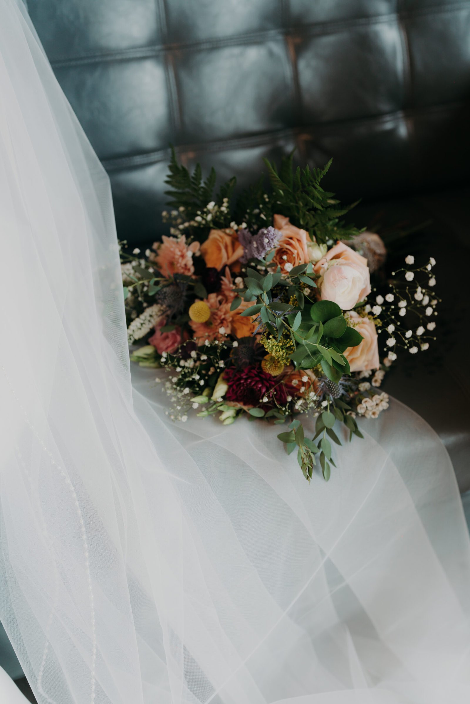 A vibrant wedding bouquet resting next to a white tulle veil on black leather couch, featuring peach roses and greenery.