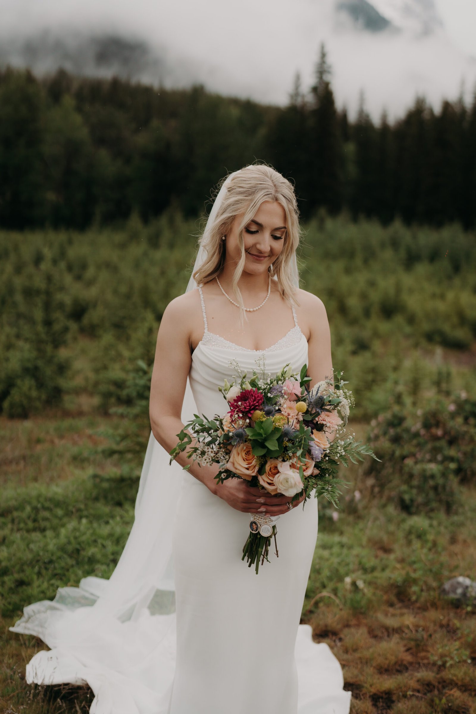 A three-quarter portrait of a bride smiling, standing in the mountain holding a rustic bouquet, her full veil billowing behind her.