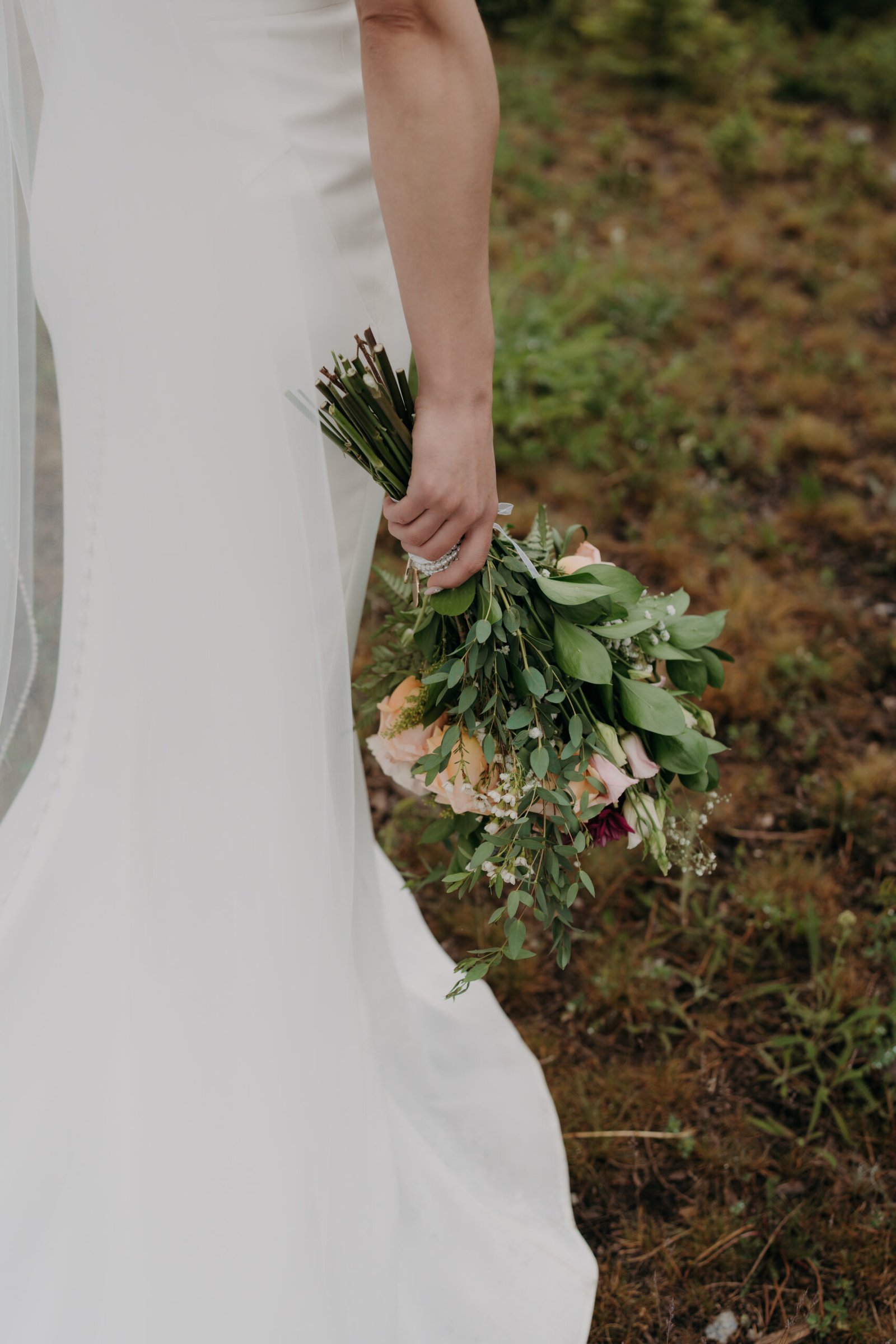 A detailed shot from behind of a bride's hand holding the wrapped stems of her wedding bouquet against white dress.