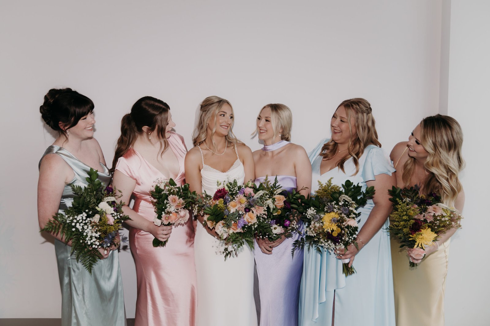 Bride in a white gown with bridesmaids in mismatched pastel dresses holding floral bouquets.
