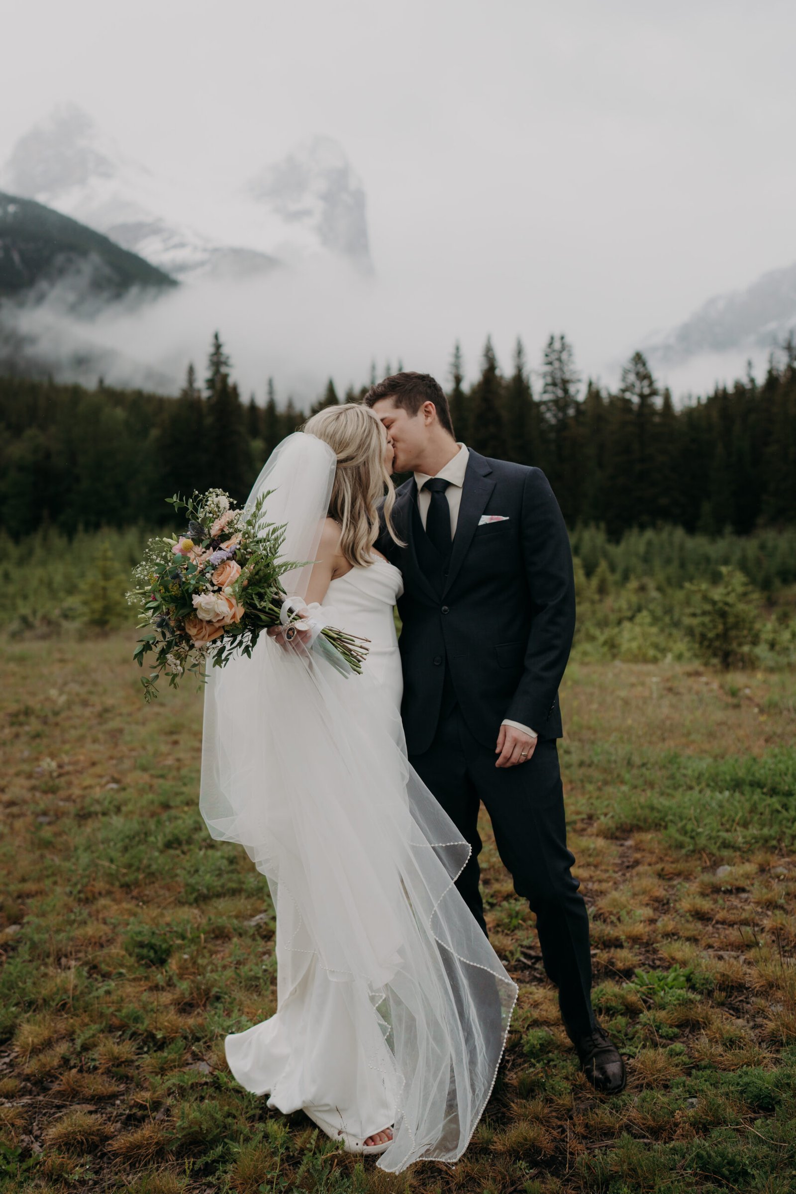 Bride and groom kissing in a misty mountain landscape with evergreen trees.